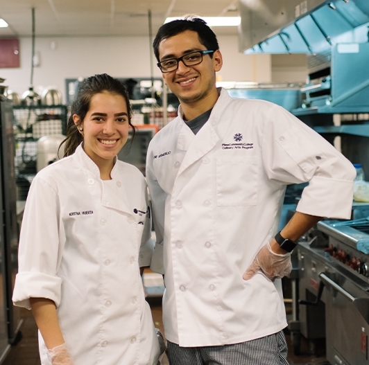 Two students stand smiling in a culinary kitchen