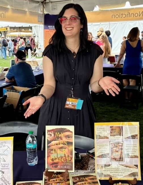 L.L. Madrid standing behind a table at an outdoor book event, smiling and gesturing toward a display of novels, bookmarks, and small decorative items, with other attendees and booths visible in the background.