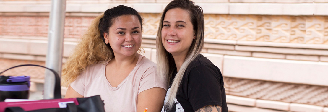 Two female students sit at a table in the courtyard of a campus, smiling at the camera