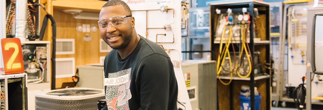 A male student sits smiling at the camera in a machinists workshop