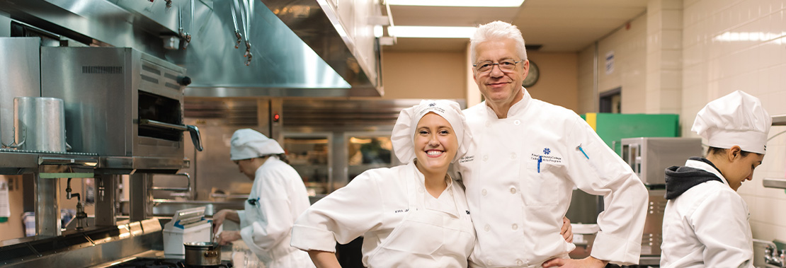 A woman and a man stand smiling in a culinary kitchen wearing culinary uniforms