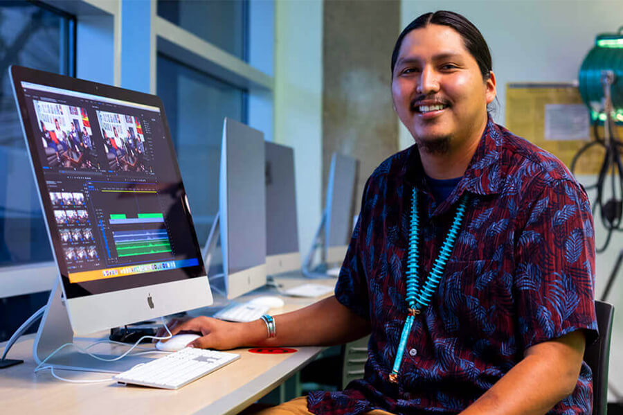 A student sits smiling while wrking on a video project in a student tech lab at Pima West Campus