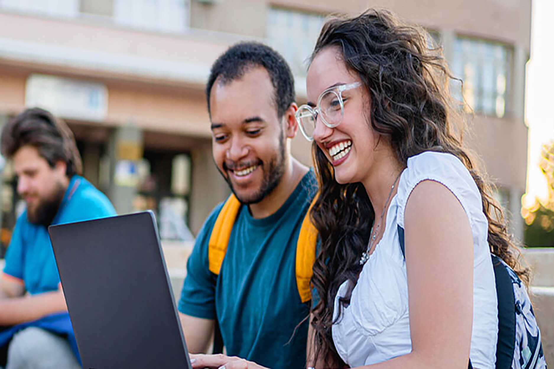 Two students sit in a campus courtyard smiling while working on a laptop 