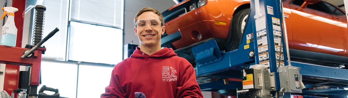 A student stands smiling in front of a vehicle on a lift