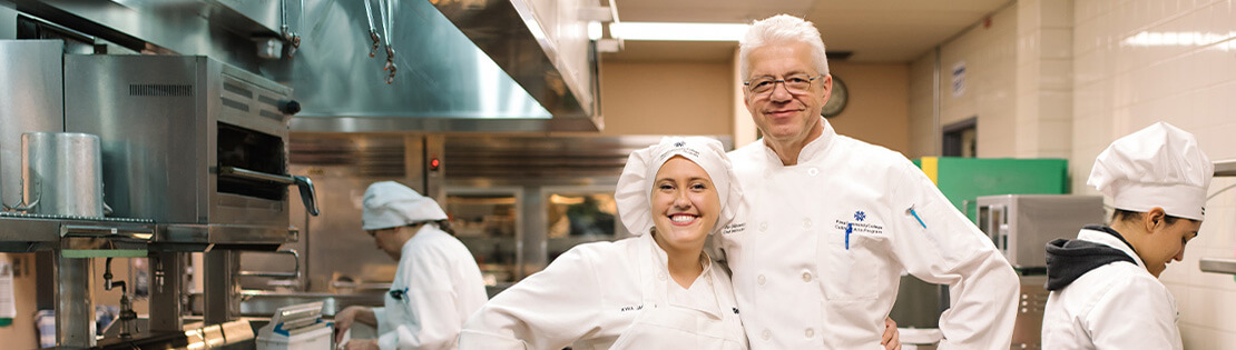 A Culinary Arts student and chef stand smiling in a Pima Culinary Kitchen