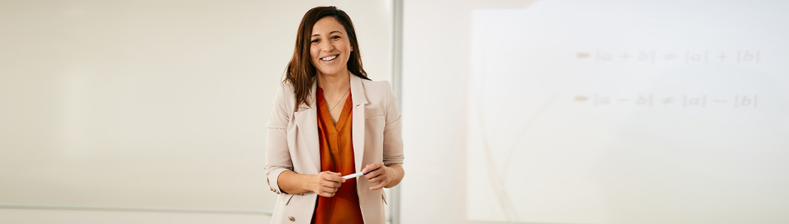 A teacher stands and smiles in front of a whiteboard