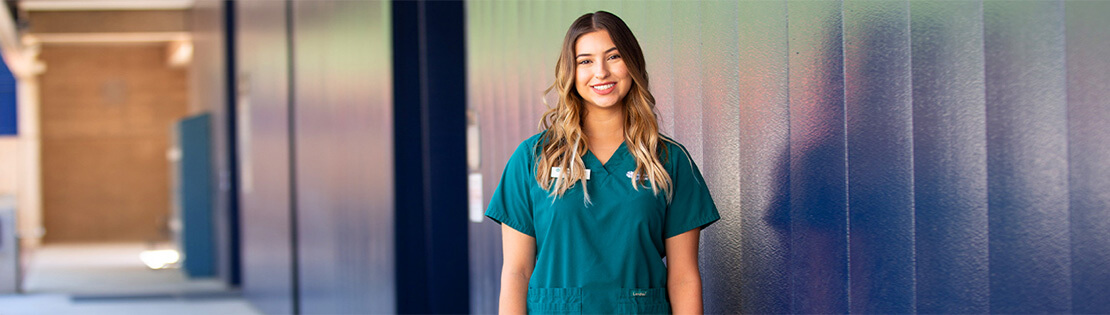 A Pima nursing student stands smiling against a wall at Pima's West Campus