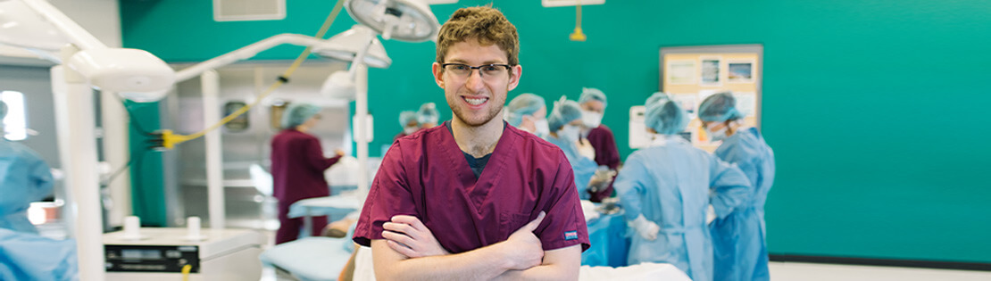 A surgical tech student poses for a photo while classmates practice behind him in a pima surgical class