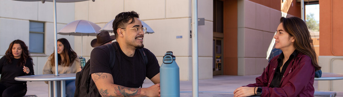 Two pima students chat at an outside table in  a courtyard at Pima's Northwest Campus