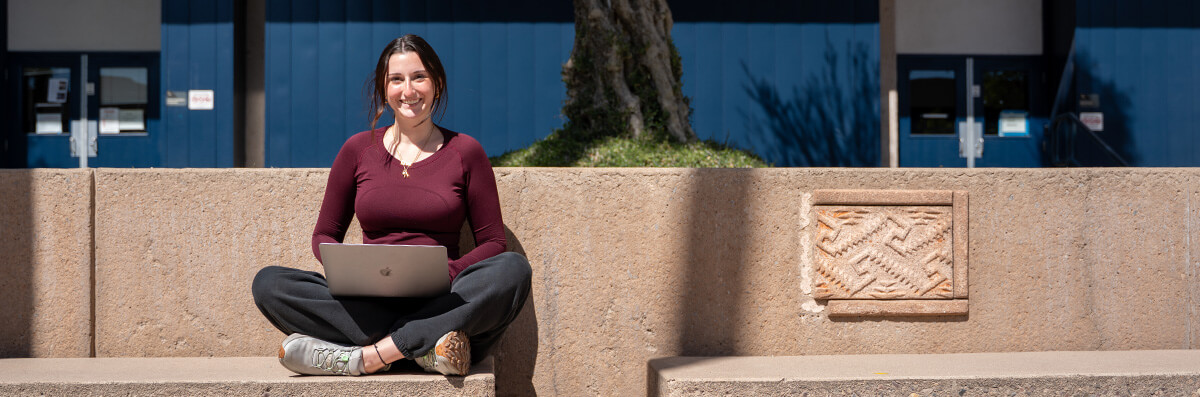 A pima student sits on her laptop smiling at West Campus.