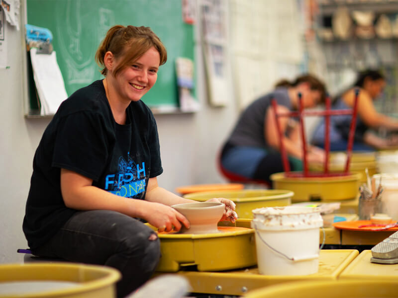 A female student works on a pottery wheel smiling at the camera